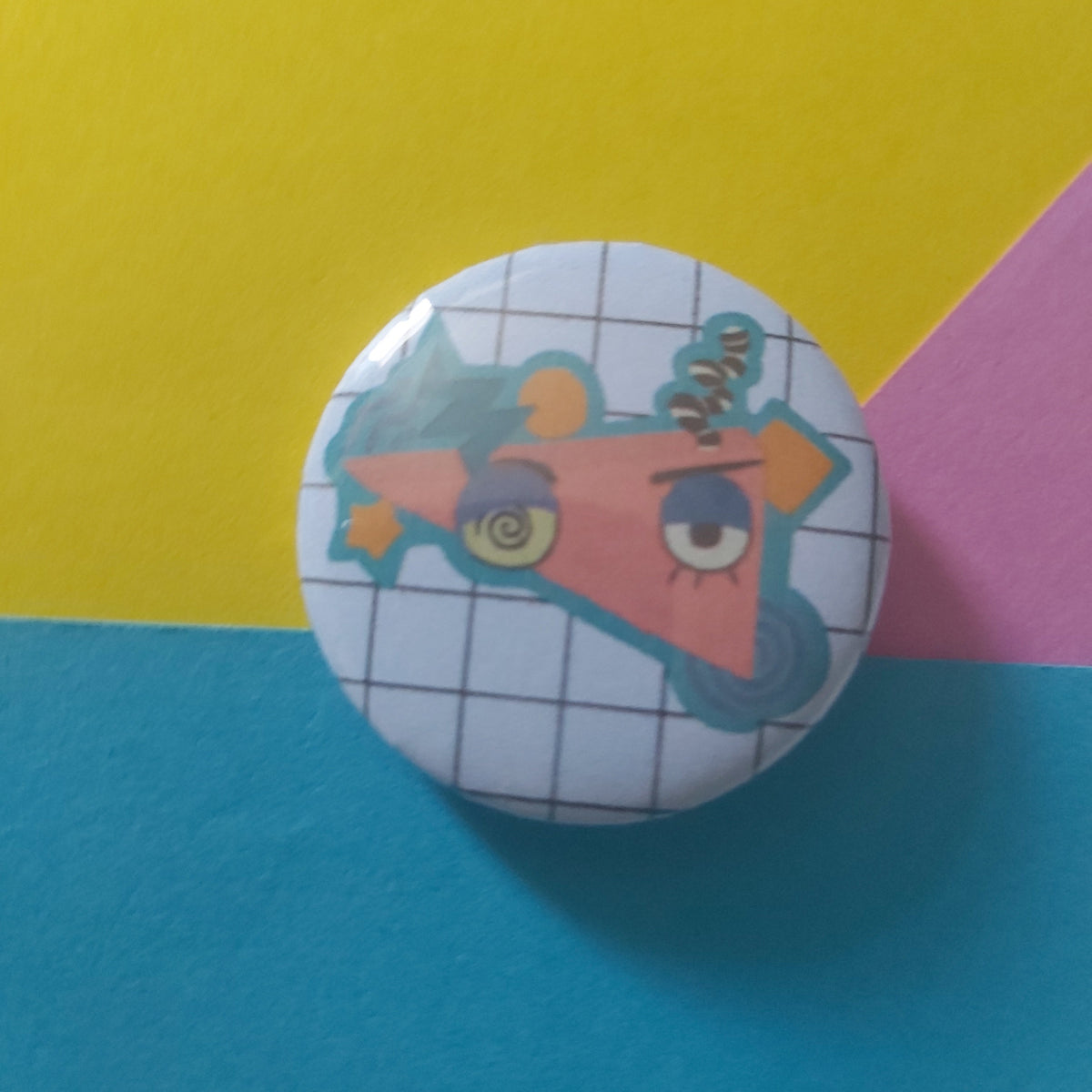 A round pinback button badge featuring a cartoon character with a spiral eye and orange hair, set against a white grid background. The button is photographed on a colorful, geometric paper backdrop split diagonally into yellow, pink, and blue sections.