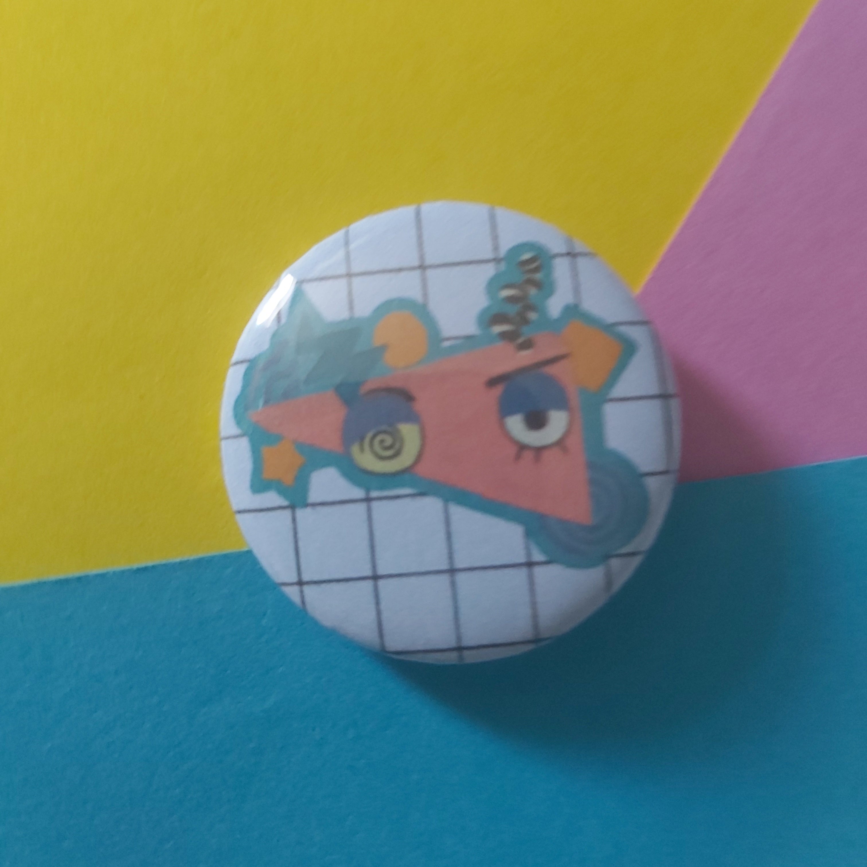 A round pinback button badge featuring a cartoon character with a spiral eye and blue outline on a white grid background, set against a colourful yellow, pink, and blue paper backdrop.