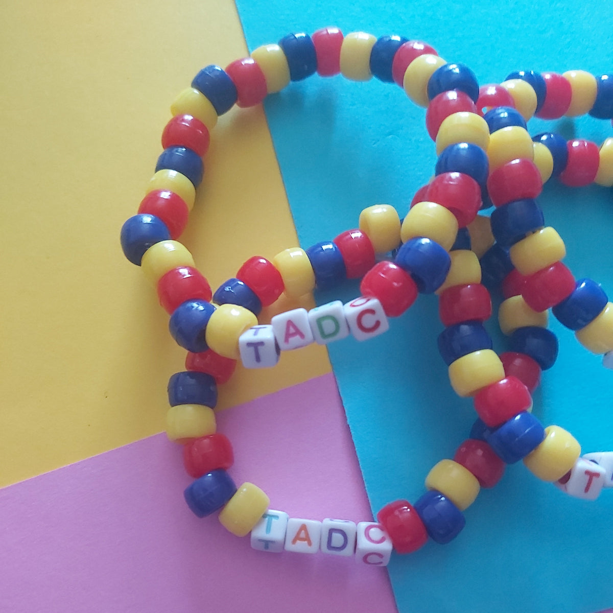 A set of colourful beaded bracelets with alternating red, yellow, and blue beads, spelt out with letter tiles reading 'TADC'. The bracelets are arranged on a background of yellow, blue, and pink paper.