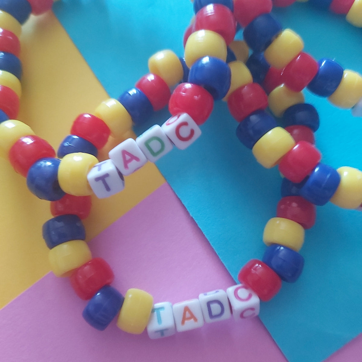A close-up of several colourful beaded bracelets arranged on pastel paper. The bracelets are made of alternating red, yellow, and blue round beads, with one bracelet featuring white letter tiles spelling out 'TADC'.