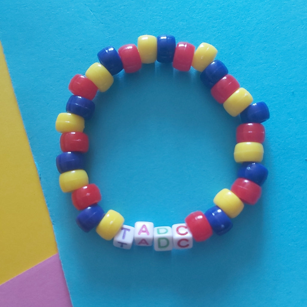 A beaded stretch bracelet with alternating red, yellow, and blue beads, featuring white letter beads spelling 'TADC' at the bottom, displayed on a bright blue background with yellow and pink paper accents.