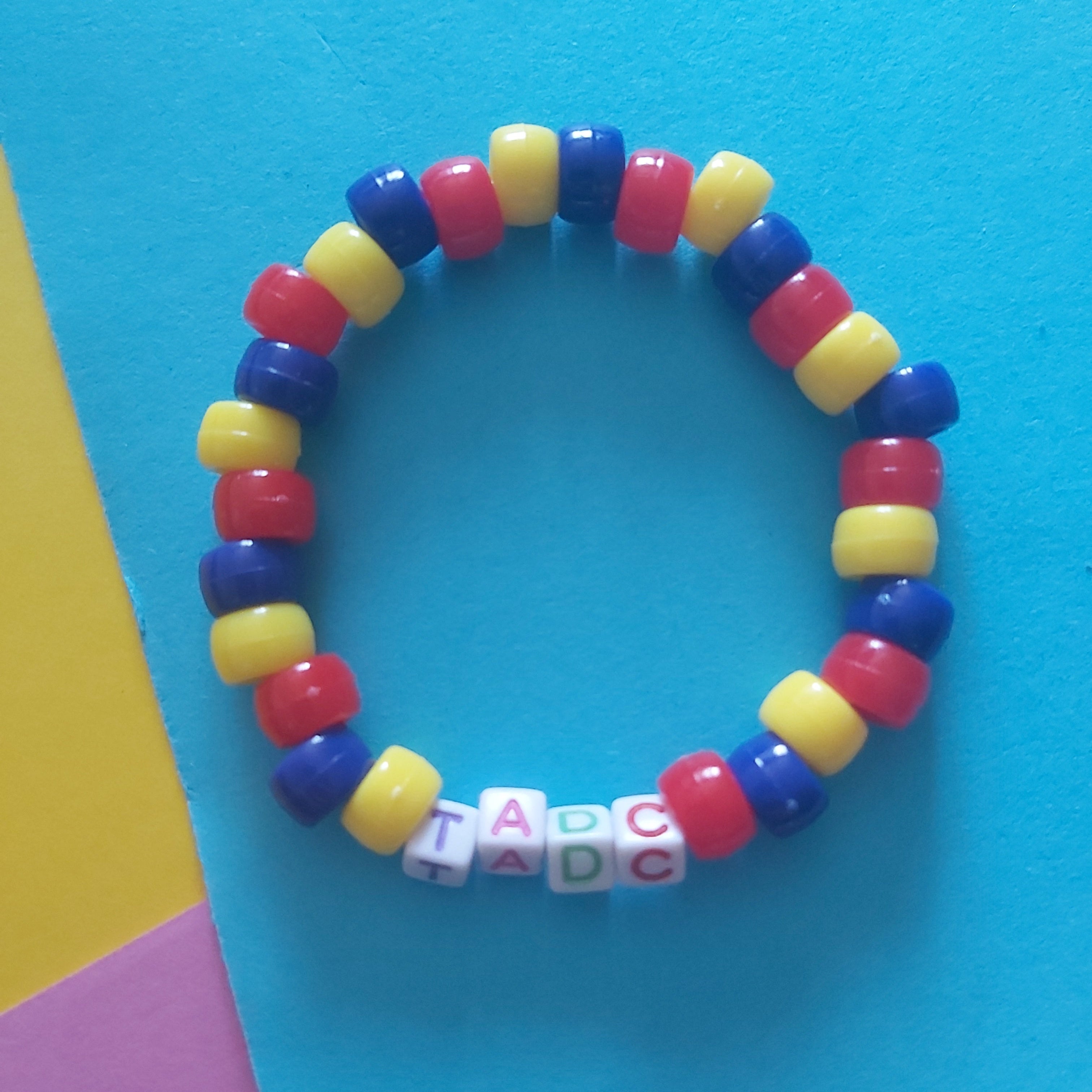 A beaded stretch bracelet with alternating red, yellow, and blue beads, featuring white letter beads spelling 'TADC' at the bottom, displayed on a bright blue background with yellow and pink paper accents.