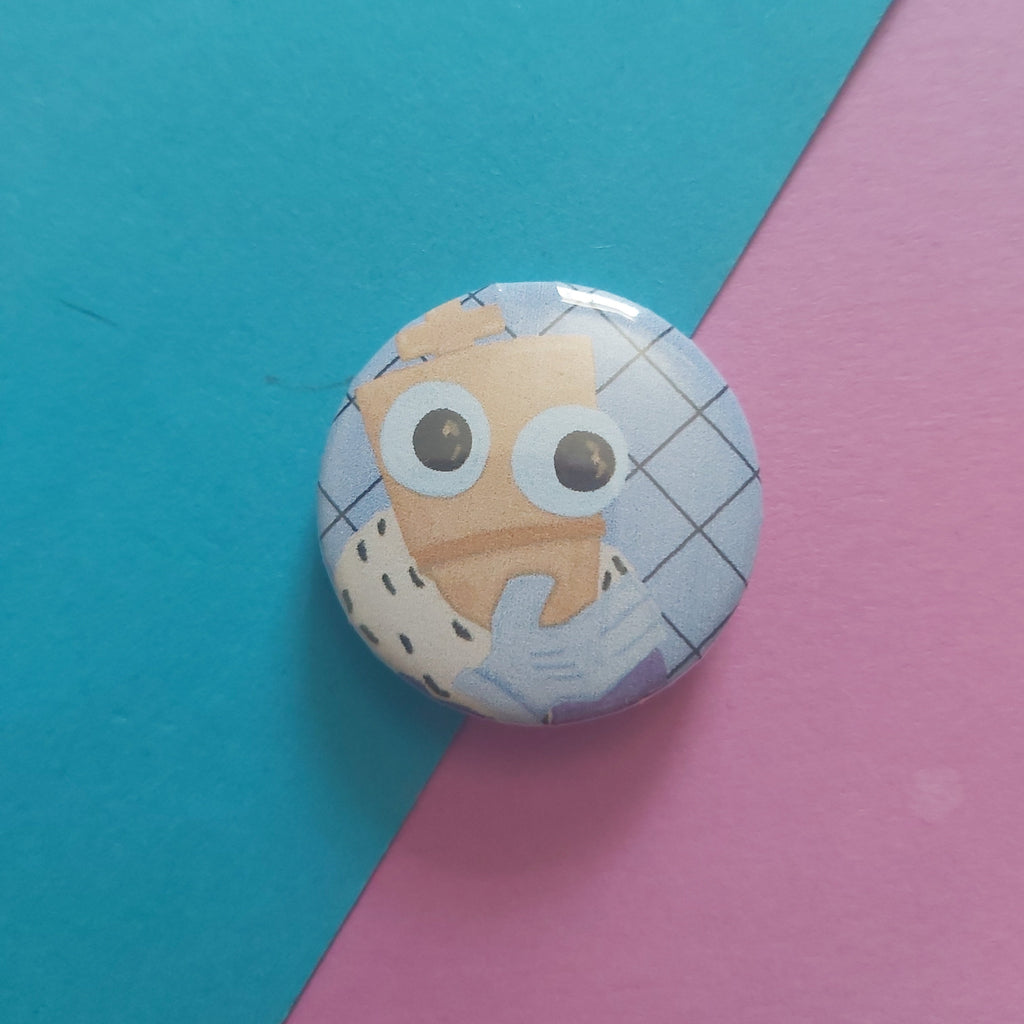 A round pinback button featuring a cartoon character with large eyes and a blue-and-white checkered pattern, photographed on a diagonally split teal and pink background.