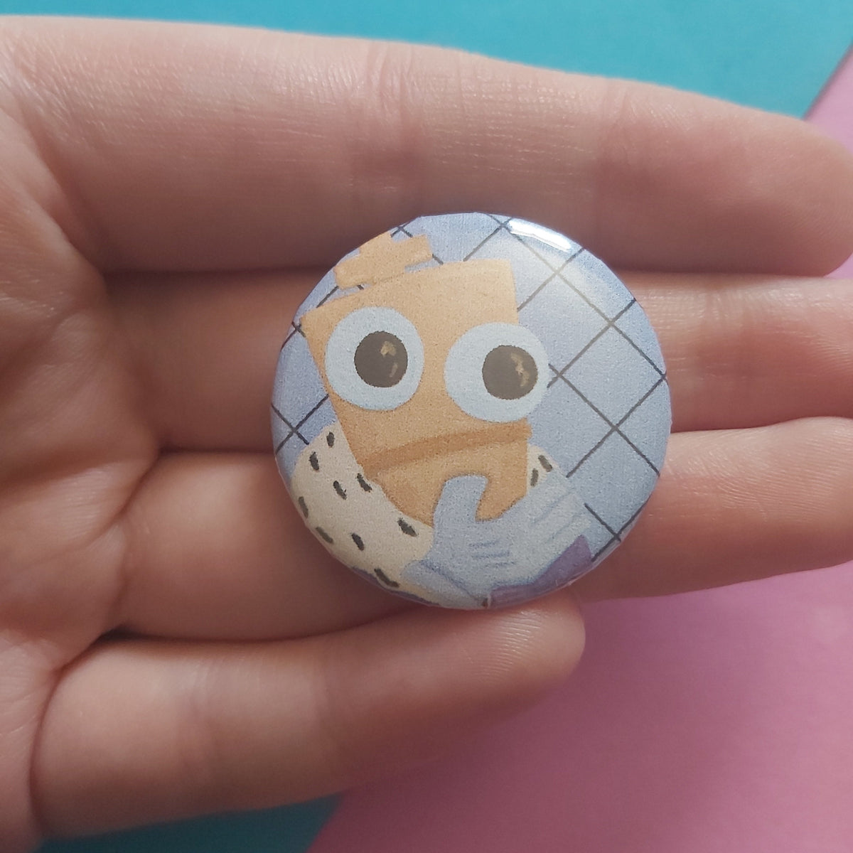 A hand holding a round pinback button badge featuring a cartoon character with large eyes against a light blue checkered background, with a pink and teal surface behind it.