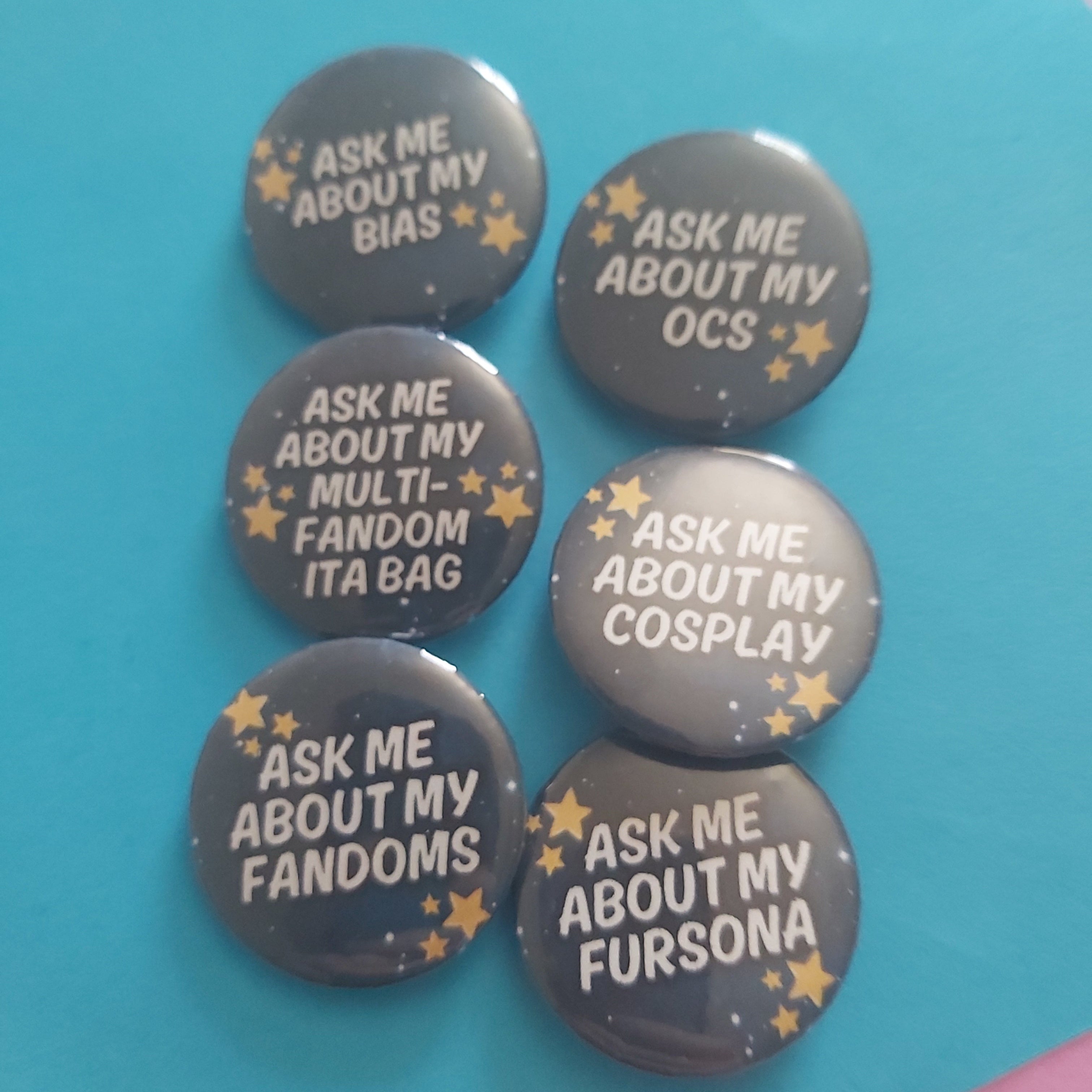 Six round pin-back buttons with dark grey backgrounds and white text reading phrases like 'ASK ME ABOUT MY BIAS', 'ASK ME ABOUT MY OCS', and 'ASK ME ABOUT MY FANDOMS', decorated with small gold stars, arranged on a bright blue background.