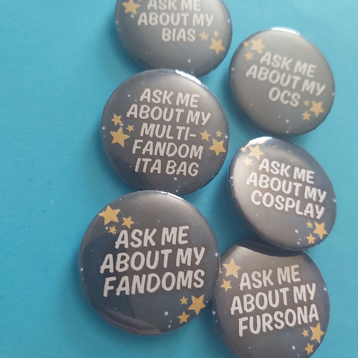 Six round pin-back buttons with a dark grey background and gold star accents, featuring humorous text about fandoms and OCS, arranged on a bright turquoise surface.