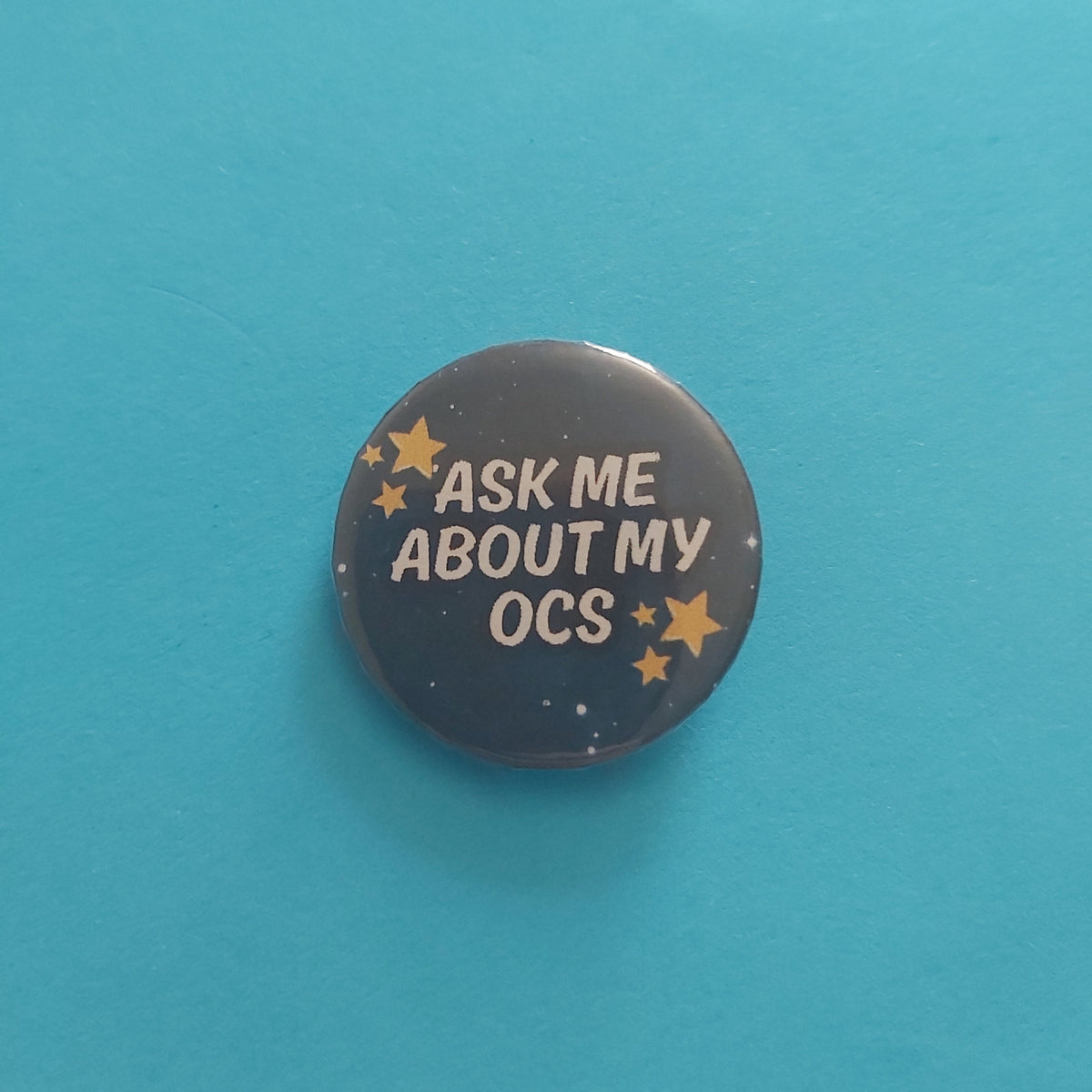 A round pinback button with a dark navy blue space-themed background, featuring the text 'ASK ME ABOUT MY OCS' in white, surrounded by small gold stars, set against a solid turquoise blue background.