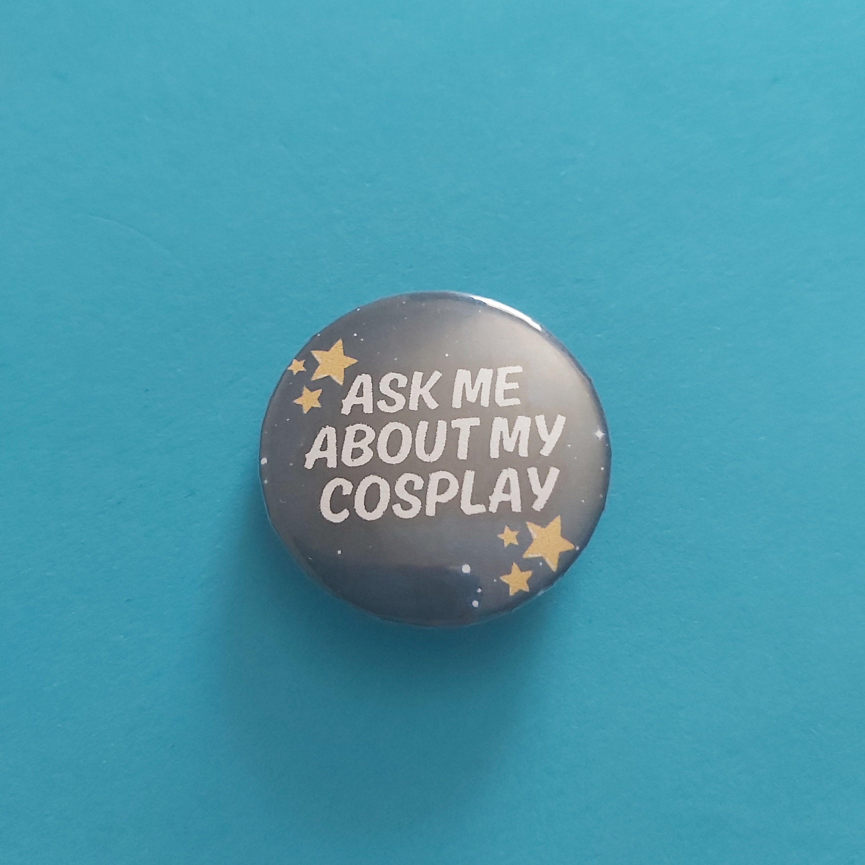 A round pinback button badge featuring a dark gray space-themed background with gold stars and the text 'ASK ME ABOUT MY COSPLAY' in white, displayed on a solid teal-blue background.