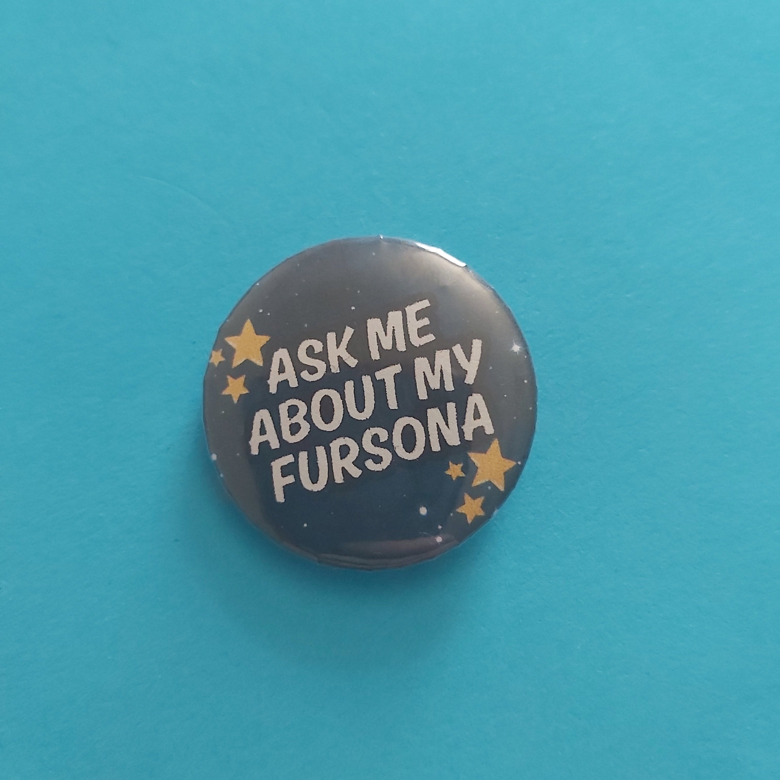A round pinback button badge featuring the text 'ASK ME ABOUT MY FURSONA' in white lettering on a dark gray starry background, accented with small gold stars, set against a solid turquoise blue background.