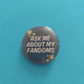 A round pinback button badge featuring the text 'ASK ME ABOUT MY FANDOMS' in white on a dark blue starry background, set against a solid turquoise blue backdrop.