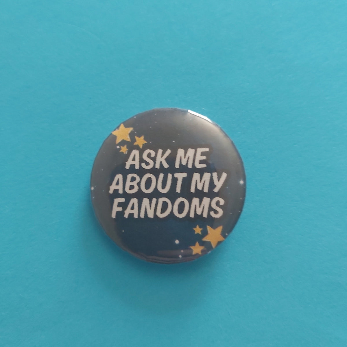 A round pinback button badge featuring the text 'ASK ME ABOUT MY FANDOMS' in white on a dark blue starry background, set against a solid turquoise blue backdrop.