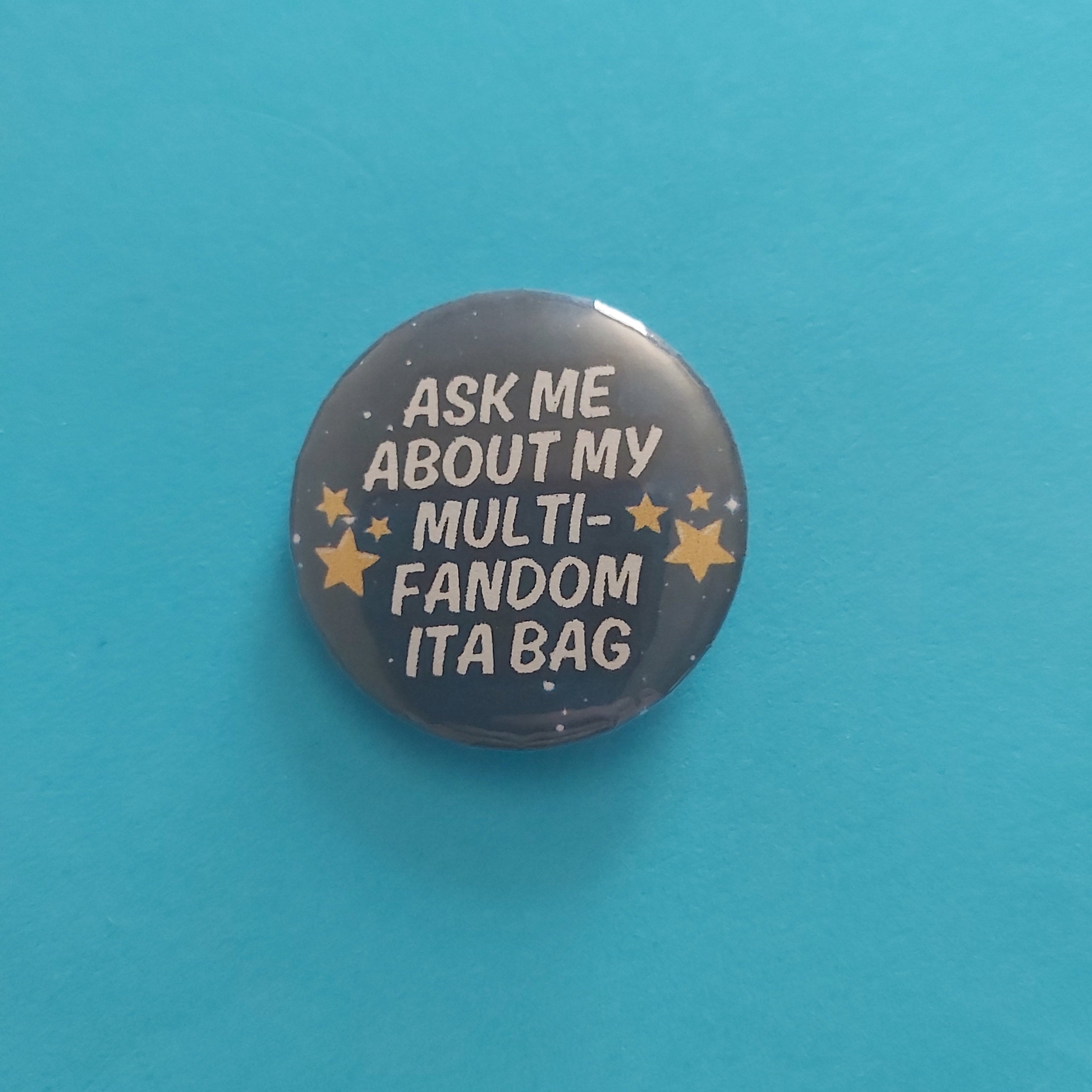 A round pinback button badge with the text 'ASK ME ABOUT MY MULTI-FANDOM ITA BAG' in white lettering, accented with small gold stars, set against a solid turquoise blue background.