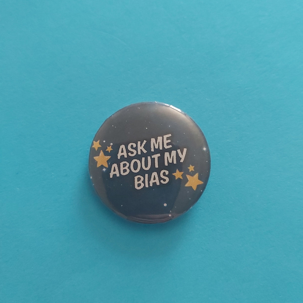 A round pinback button with the text 'ASK ME ABOUT MY BIAS' in white, surrounded by small gold stars on a dark background, placed on a solid turquoise surface.