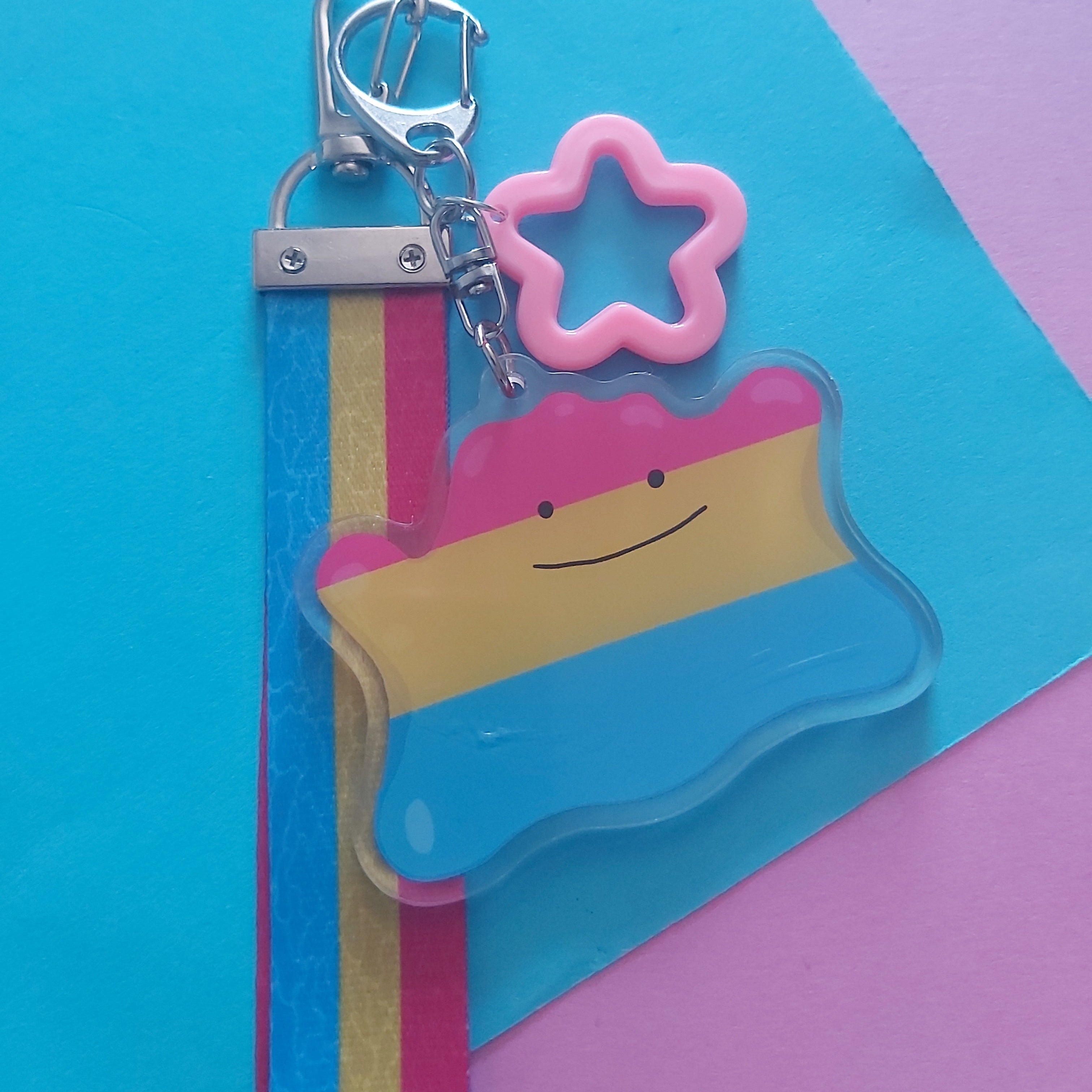 A keychain featuring a translucent star-shaped charm with a pink outline, attached to a silver lobster clasp. The charm is decorated with horizontal stripes of pink, yellow, and blue, and is displayed on a colourful background of blue, pink, and yellow paper.