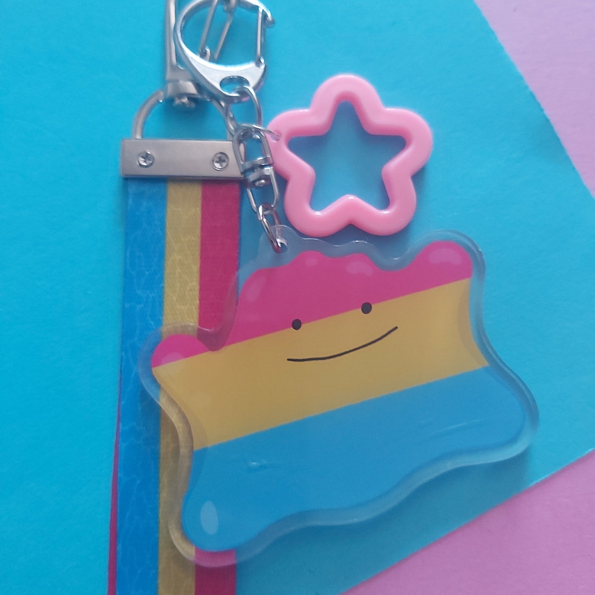 A keychain featuring a charm with a pink, yellow, and blue horizontal stripe pattern and a simple smiley face, attached to a silver lobster clasp and a pink star-shaped charm, set against a colourful geometric background.