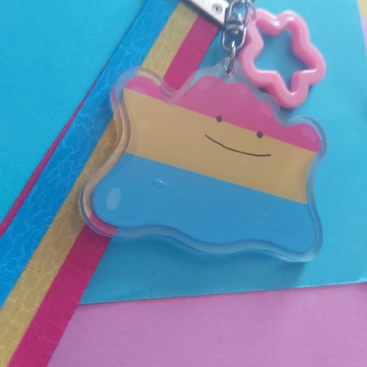 A shaped acrylic keychain charm with a smiling face, attached to a silver clasp and a pink star-shaped ring, shown on a colourful background of blue, pink, and gold paper.