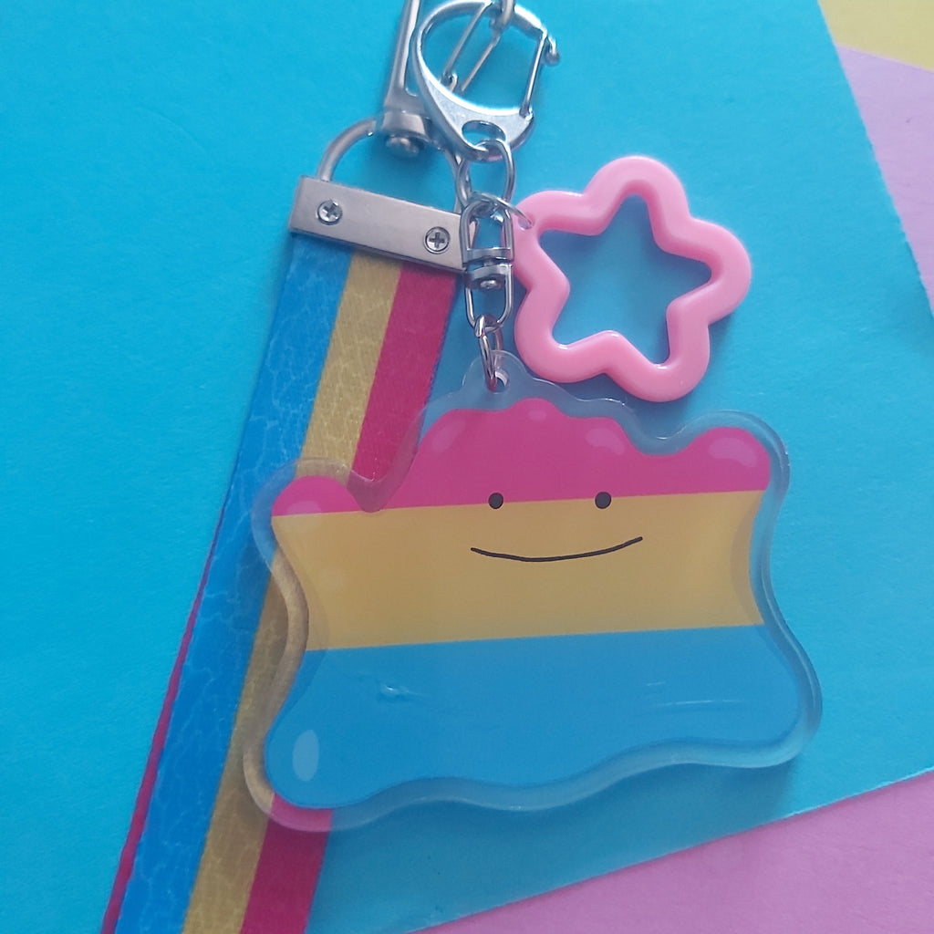 An acrylic keychain featuring a smiling character with pink, yellow, and blue horizontal stripes, a pink star charm, and a silver metal clasp, displayed on a colourful background of blue, pink, and yellow paper.