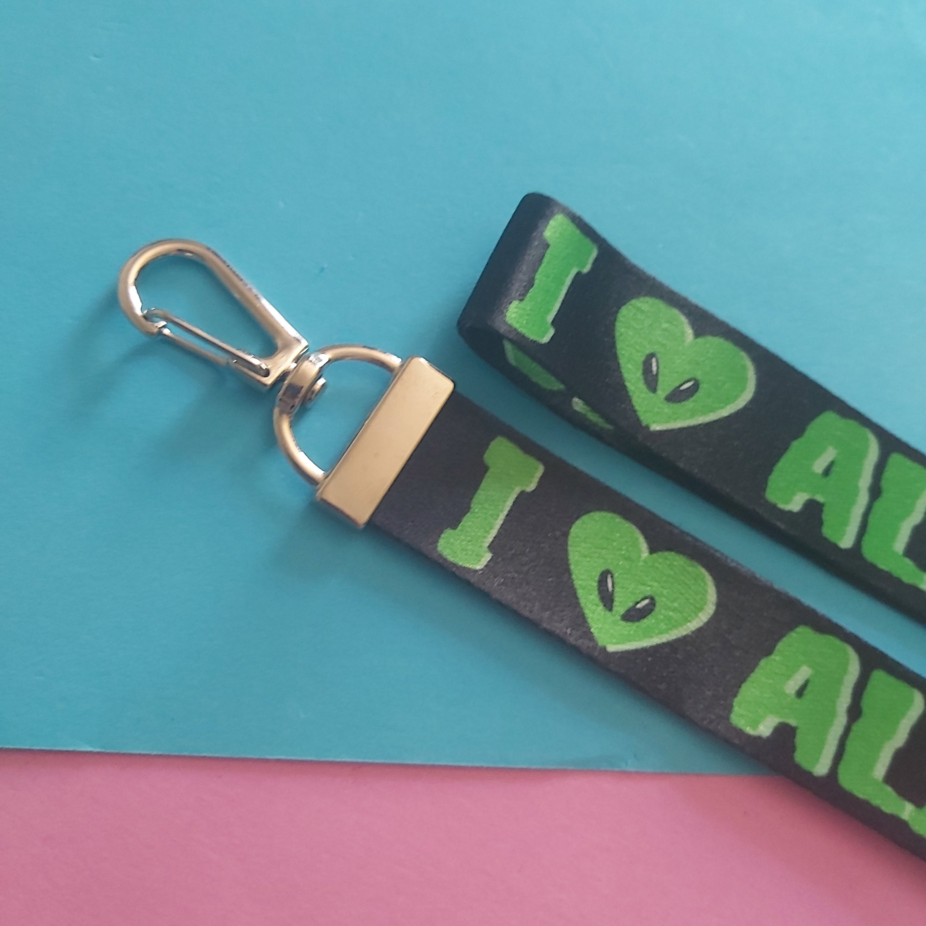A black fabric wristlet keychain strap with a silver metal clasp, featuring a green graphic that reads 'I heart Aliens' with alien eyes, displayed on a split turquoise and pink background.