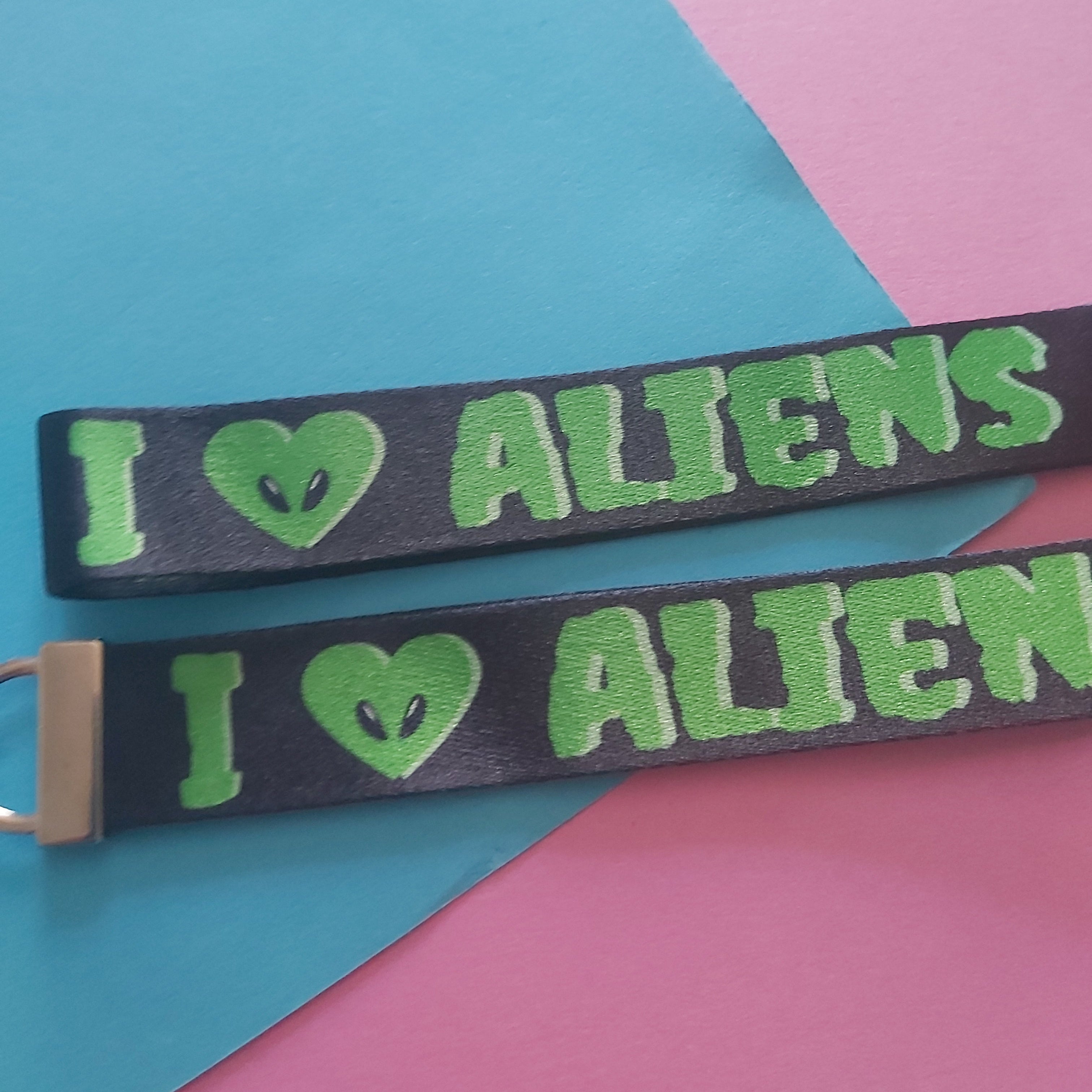 Two black woven straps with green embroidered text reading 'I [alien heart] ALIENS' laid diagonally on a colorful background of blue, pink, and yellow paper.