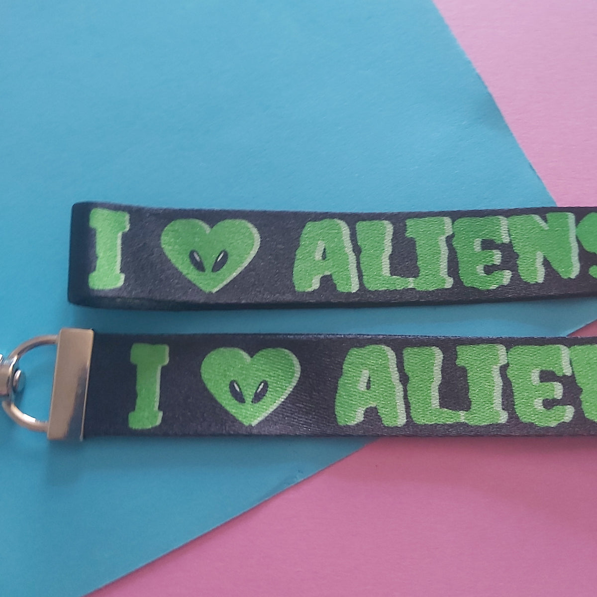 A black wrist strap keychain with green text reading 'I [alien heart] ALIENS' and a silver metal keyring, displayed on a colorful background of blue, pink, and yellow paper.