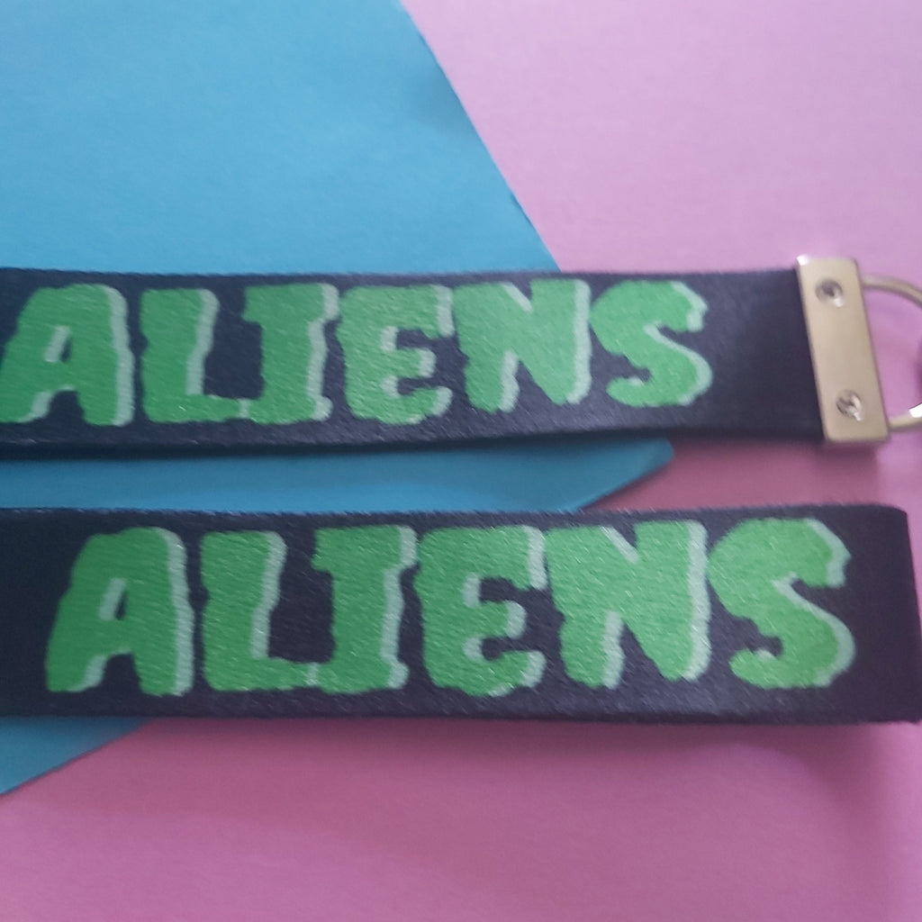 Two black fabric keychains with green 'I Heart ALIENS' text, displayed on a colourful background of pink, blue, and yellow paper.