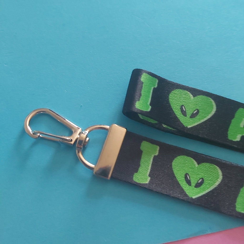 A close-up of a black wristlet keychain strap with green alien-themed graphics and a silver metal clasp, placed on a bright blue surface with a pink corner visible.
