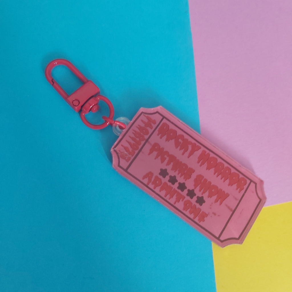 A pink acrylic keychain shaped like a ticket, attached to a red carabiner clip, set against a colorful background of cyan, pink, and yellow blocks.