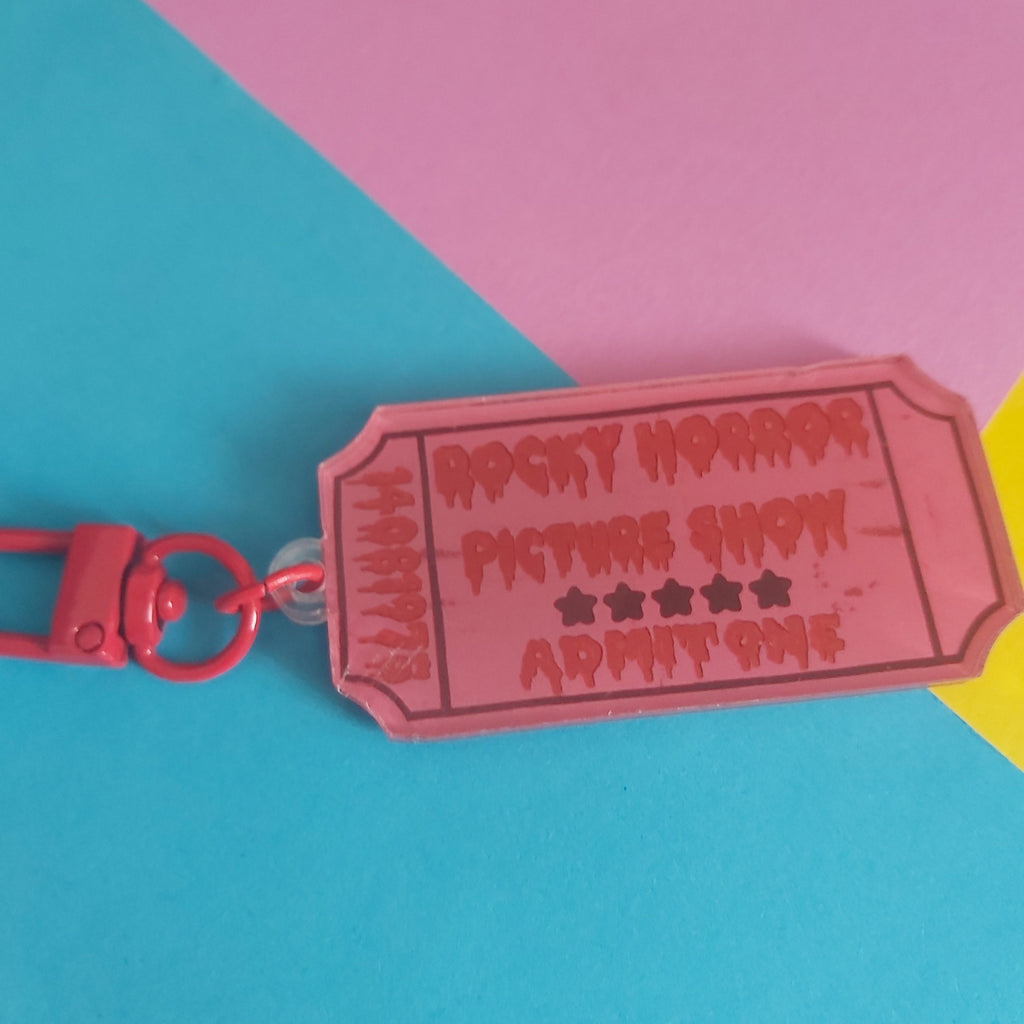 A pink acrylic keychain shaped like a retro movie ticket with 'Rocky Horror Picture Show' and 'Admit One' printed in red, attached to a red key ring, set against a colourful pink, blue, and yellow background.