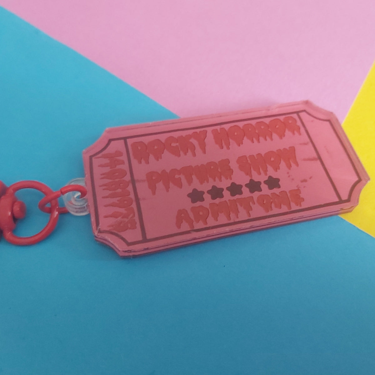 A pink ticket-shaped keychain with 'Rocky Horror Picture Show Admit One' on it, attached to a red key ring and clasp, set against a colourful geometric background of pink, yellow, and blue paper.
