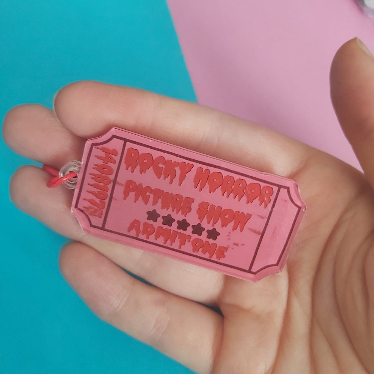 A hand holding a pink ticket-shaped keychain charm that reads 'Rocky Horror Picture Show' with star icons, against a background of pink and teal paper.