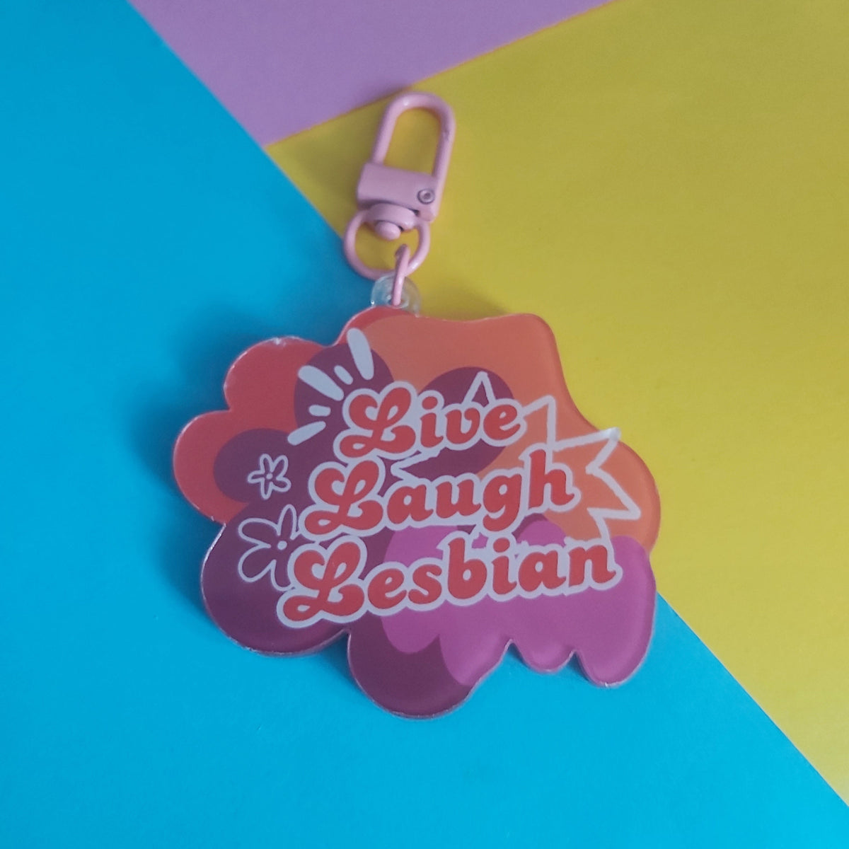 A pink and orange cloud-shaped keychain with the text 'Live Laugh Lesbian' in stylised lettering, shown on a diagonally split blue, yellow, and pink background.