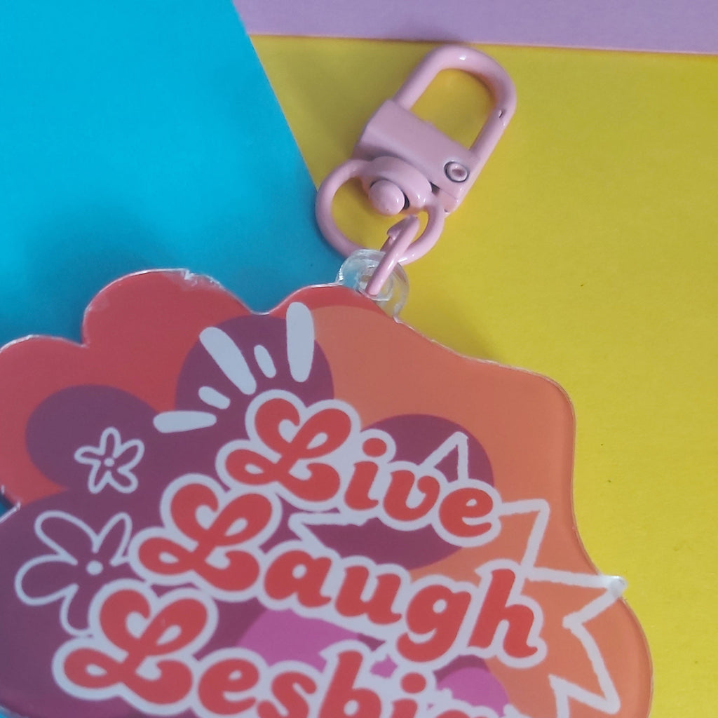 A pink and orange cloud-shaped acrylic keychain with the words 'Live Laugh Lesbian' in orange bubble letters, shown on a bright blue and yellow background.
