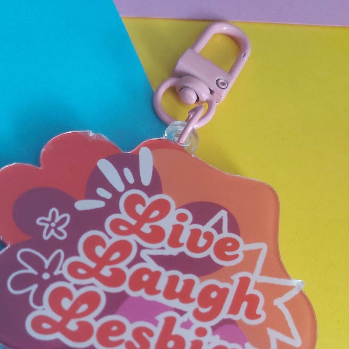 A pink and orange cloud-shaped acrylic keychain with the words 'Live Laugh Lesbian' in orange bubble letters, shown on a bright blue and yellow background.