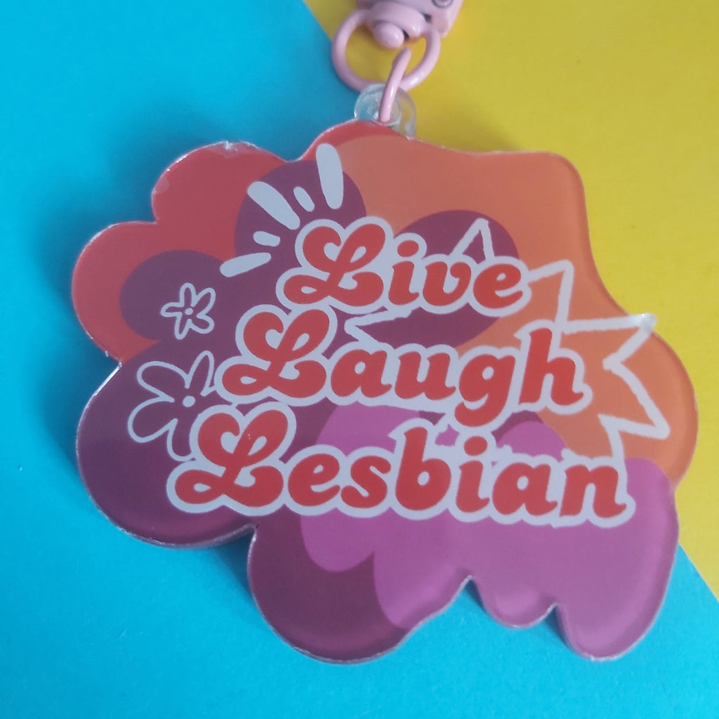 A colourful acrylic keychain with a pink lobster clasp, featuring the text 'Live Laugh Lesbian' in orange script over a pink and orange cloud-like design, set against a split blue and yellow background.
