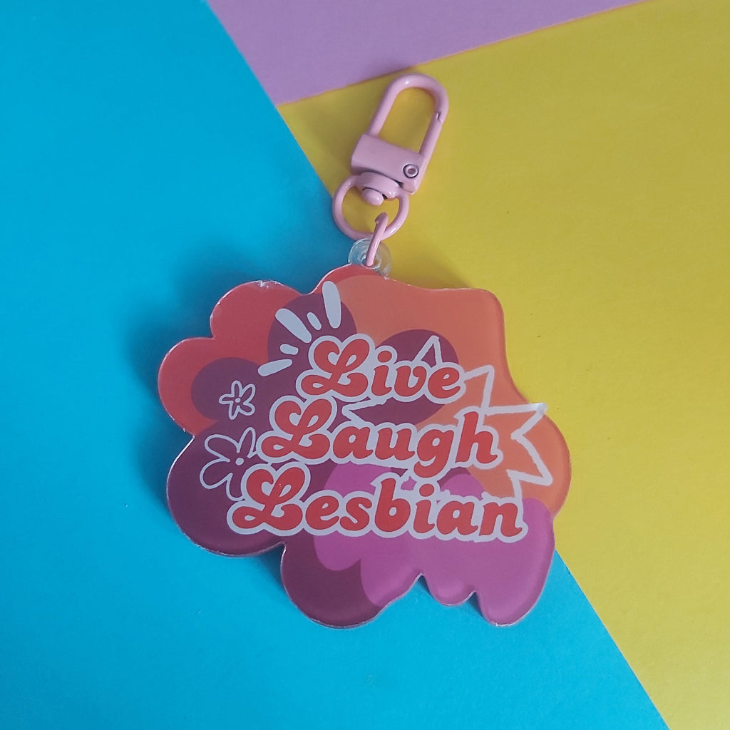 A pink and orange acrylic keychain with the text 'Live Laugh Lesbian' in stylised lettering, attached to a light pink lobster clasp, set against a diagonally split blue, yellow, and purple background.