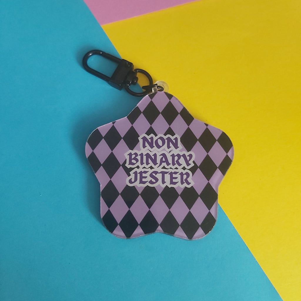 A star-shaped keychain with a black and lavender harlequin pattern and the text 'NON BINARY JESTER' in purple, featuring a black carabiner clip, set against a colorful background of pink, yellow, and blue paper.