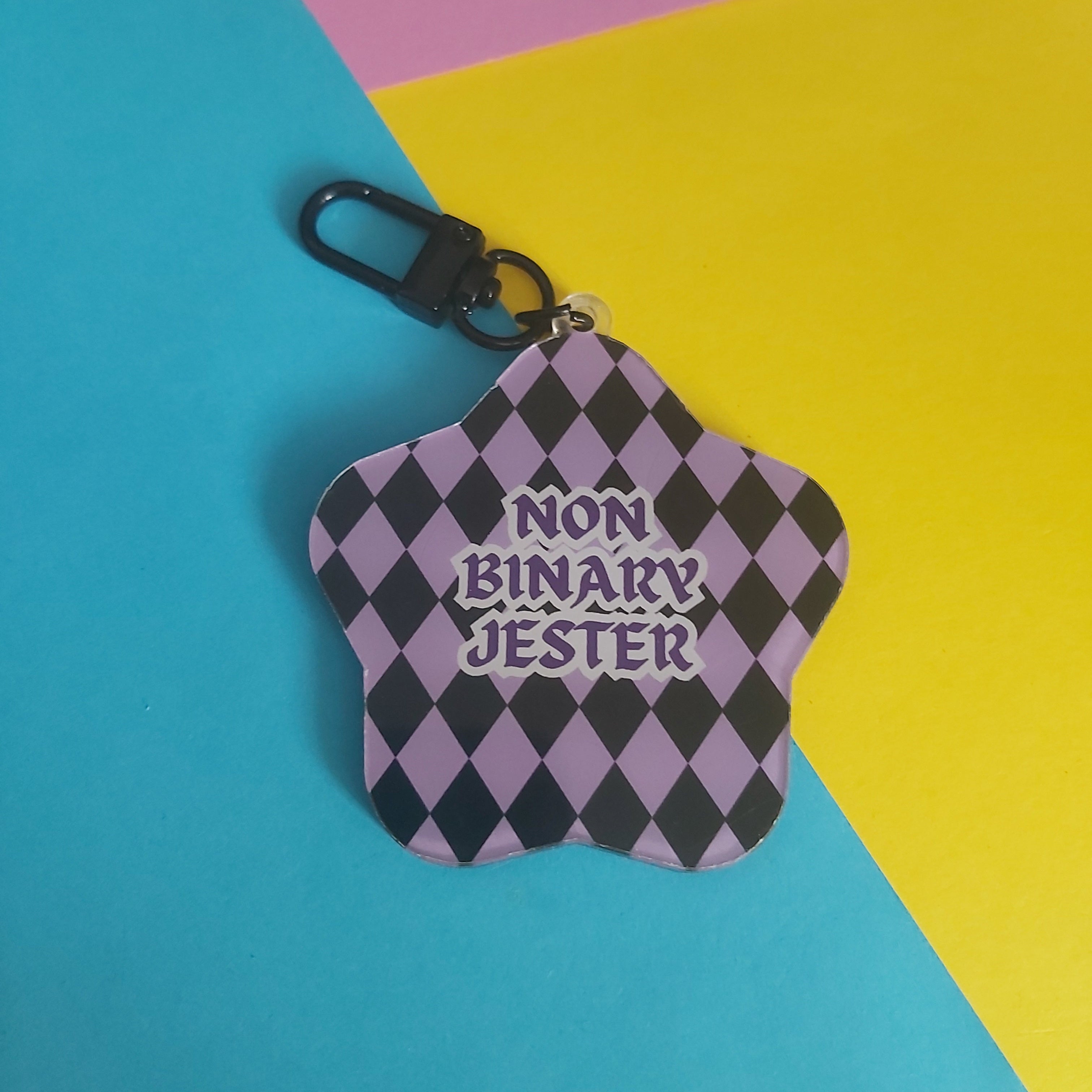 A star-shaped keychain with a black and lavender harlequin pattern and the text 'NON BINARY JESTER' in purple, featuring a black carabiner clip, set against a colorful background of pink, yellow, and blue paper.