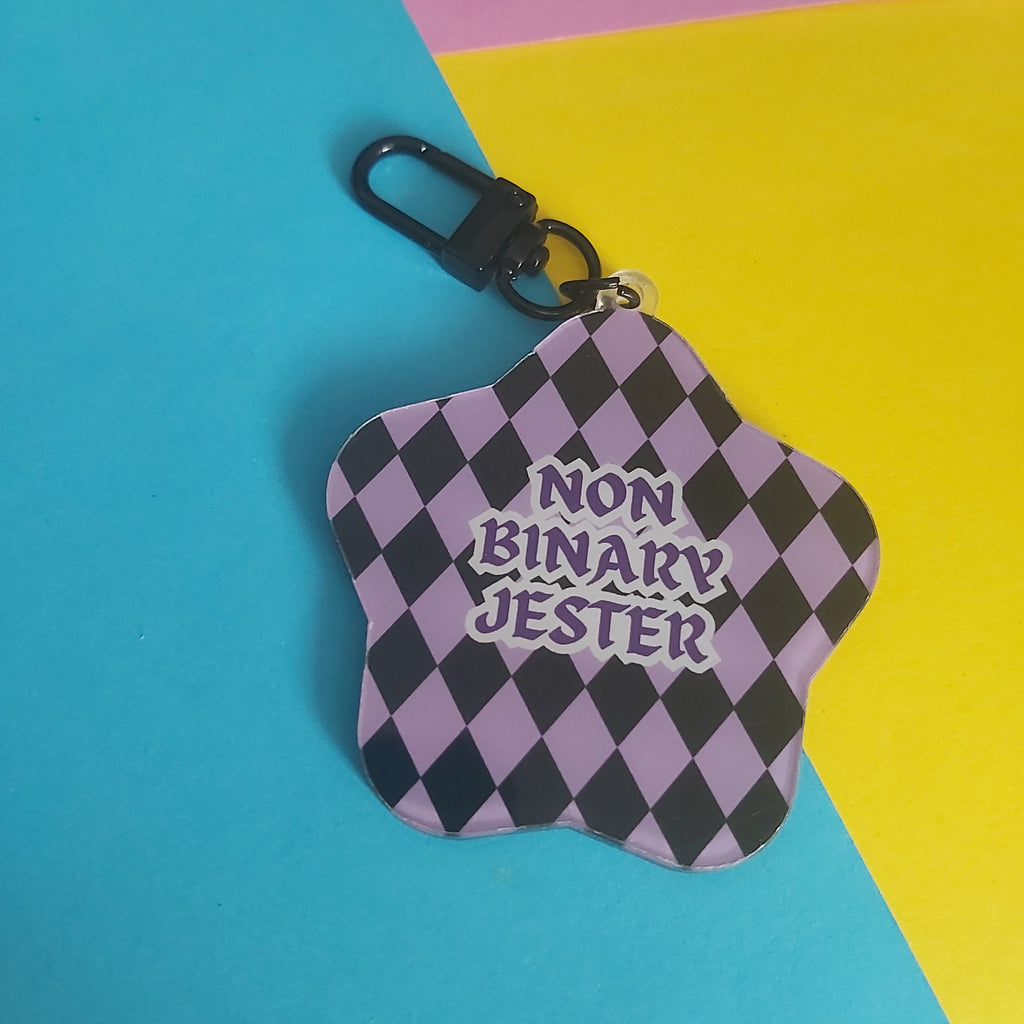 A star-shaped keychain with a black and lavender-purple checkered pattern and the text 'NON BINARY JESTER' in purple, attached to a black carabiner clip, set against a colorful background of blue, yellow, and pink paper.