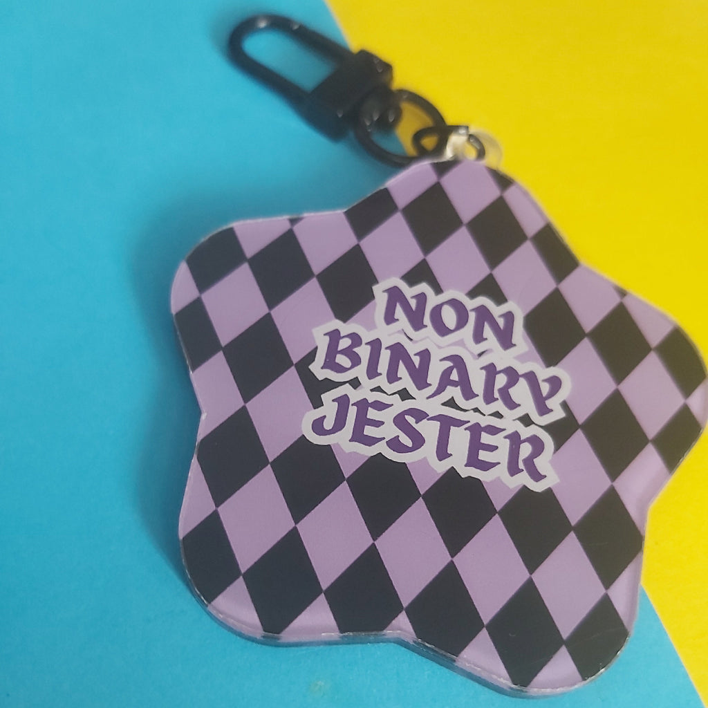 A star-shaped acrylic keychain with a black and lavender checkered pattern and the text 'NON BINARY JESTER' in purple, shown with a black carabiner clip on a split blue and yellow background.