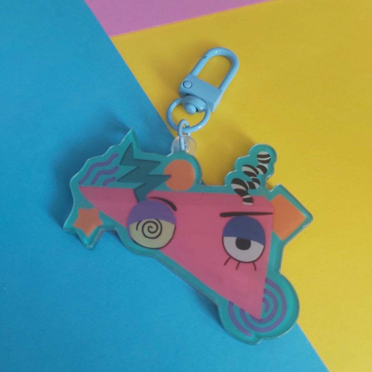 A colourful acrylic keychain charm featuring a stylized cartoon face inside a pink triangle, attached to a light blue lobster clasp, set against a diagonally split pink, yellow, and blue background.
