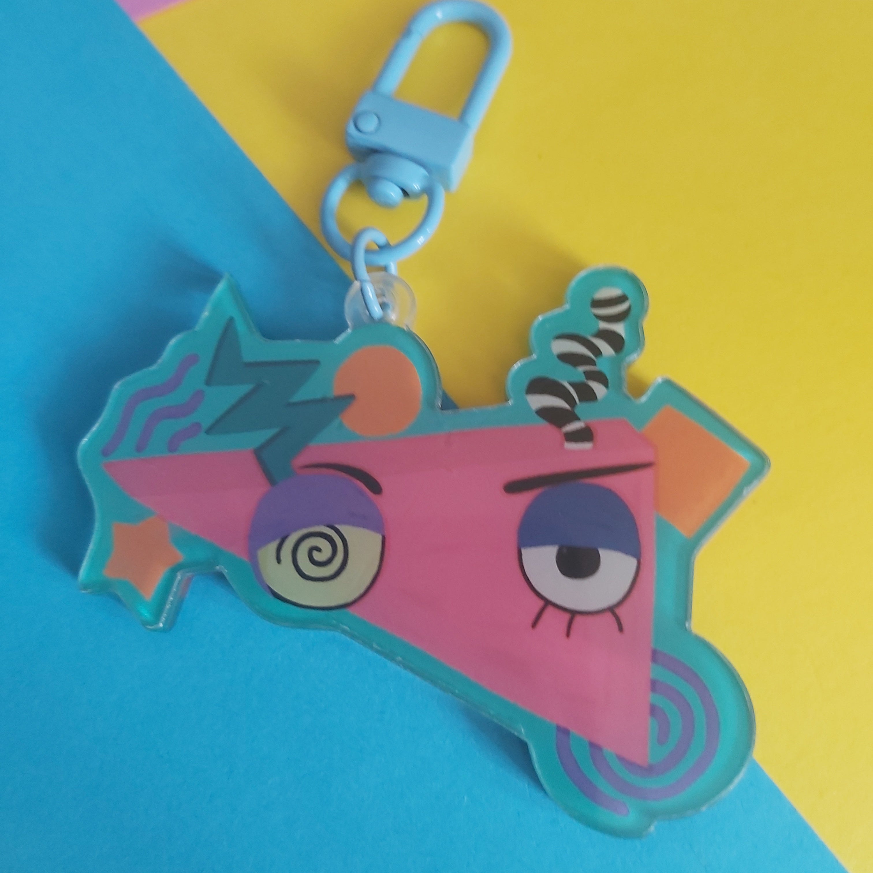 A colourful acrylic keychain charm featuring a pink triangular face with cartoon eyes, purple eyebrows, and a light blue clasp, displayed on a diagonally split yellow and blue background.