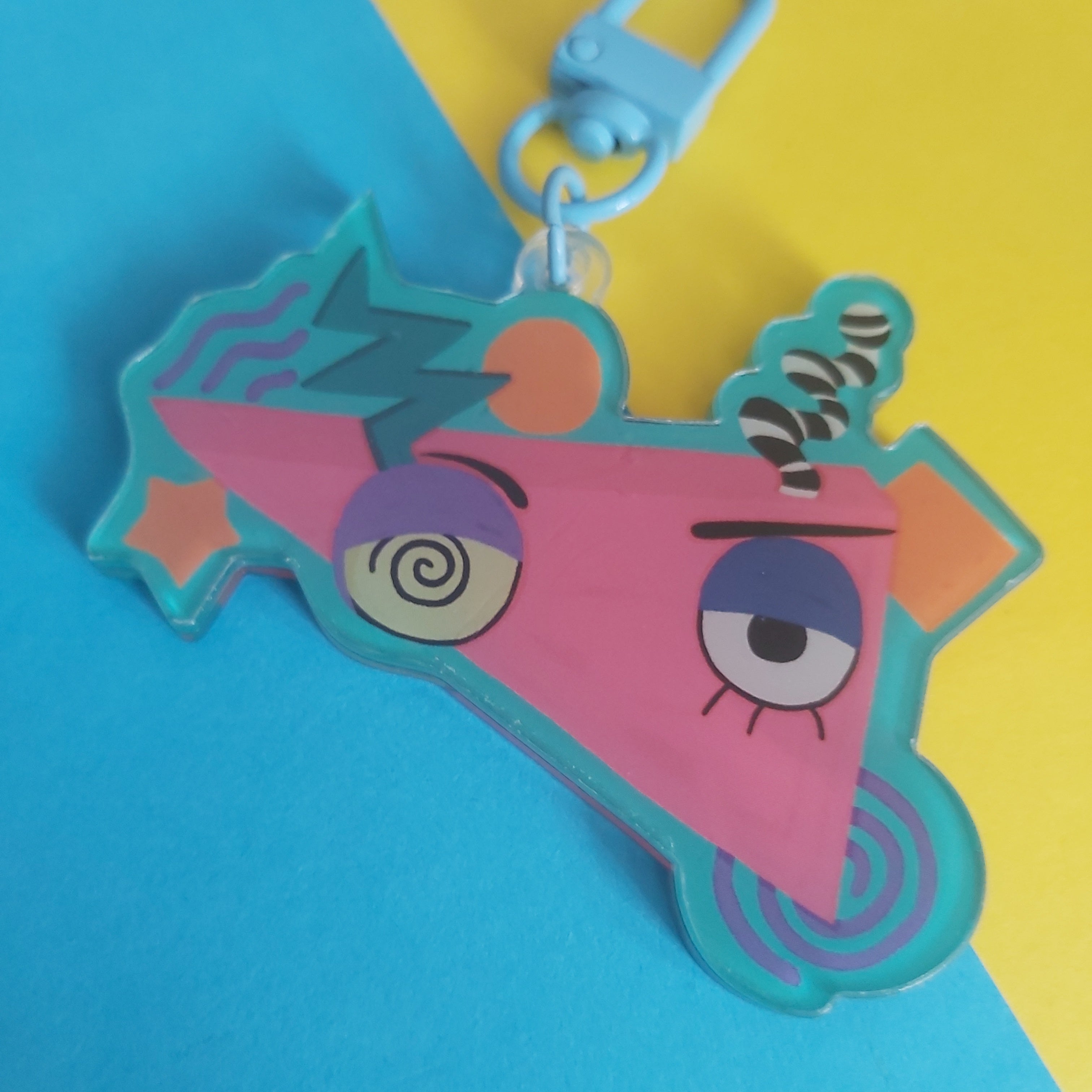 A colourful acrylic keychain featuring a cartoon triangle character with a pink body, blue outlines, and expressive eyes, attached to a light blue clasp, set against a diagonally split blue and yellow background.