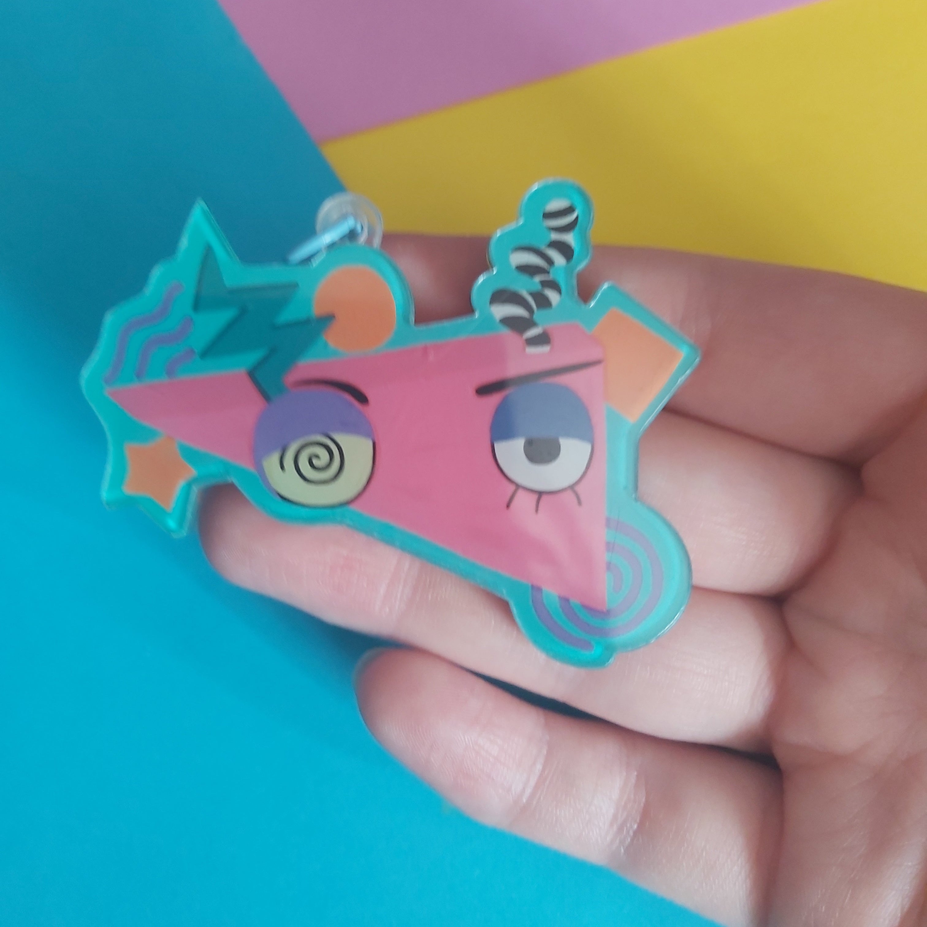A hand holding a colourful, cartoonish triangular keychain charm with a pink face, blue outlines, and a spiral horn, set against a background of bright blue, pink, and yellow paper.