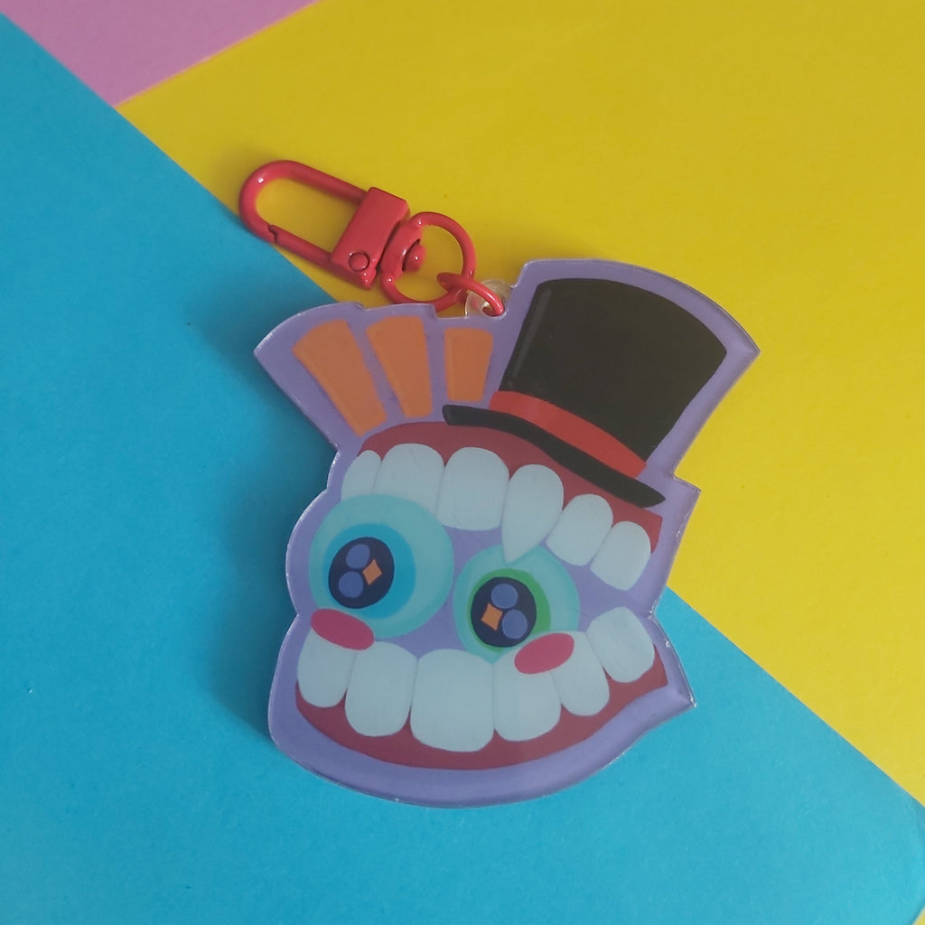 A colorful acrylic keychain featuring a stylized cartoon monster face with large blue eyes, white teeth, a black top hat, and a red carabiner clasp, set against a diagonally split yellow and blue background.