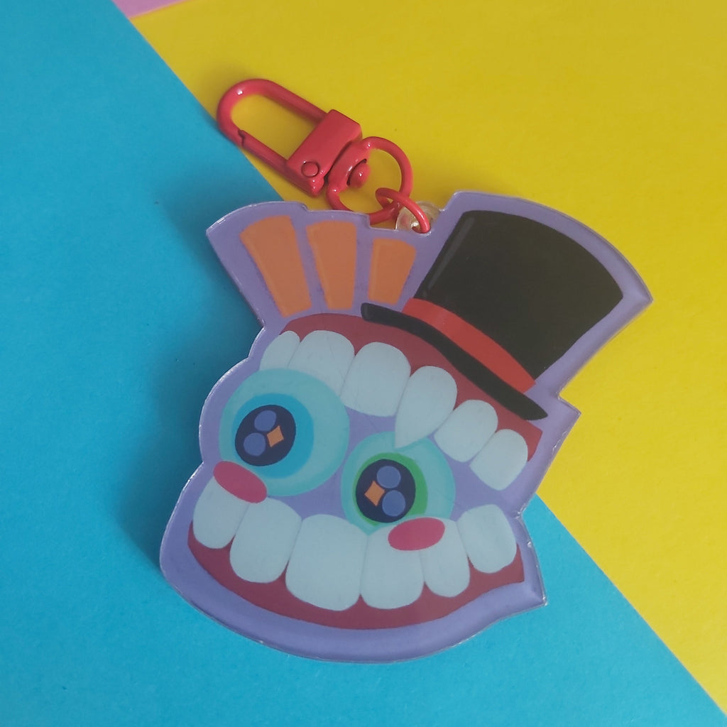 A colorful acrylic keychain charm shaped like a smiling monster face with large eyes, white teeth, pink cheeks, a black top hat, and a red clasp, photographed on a bright blue and yellow background.