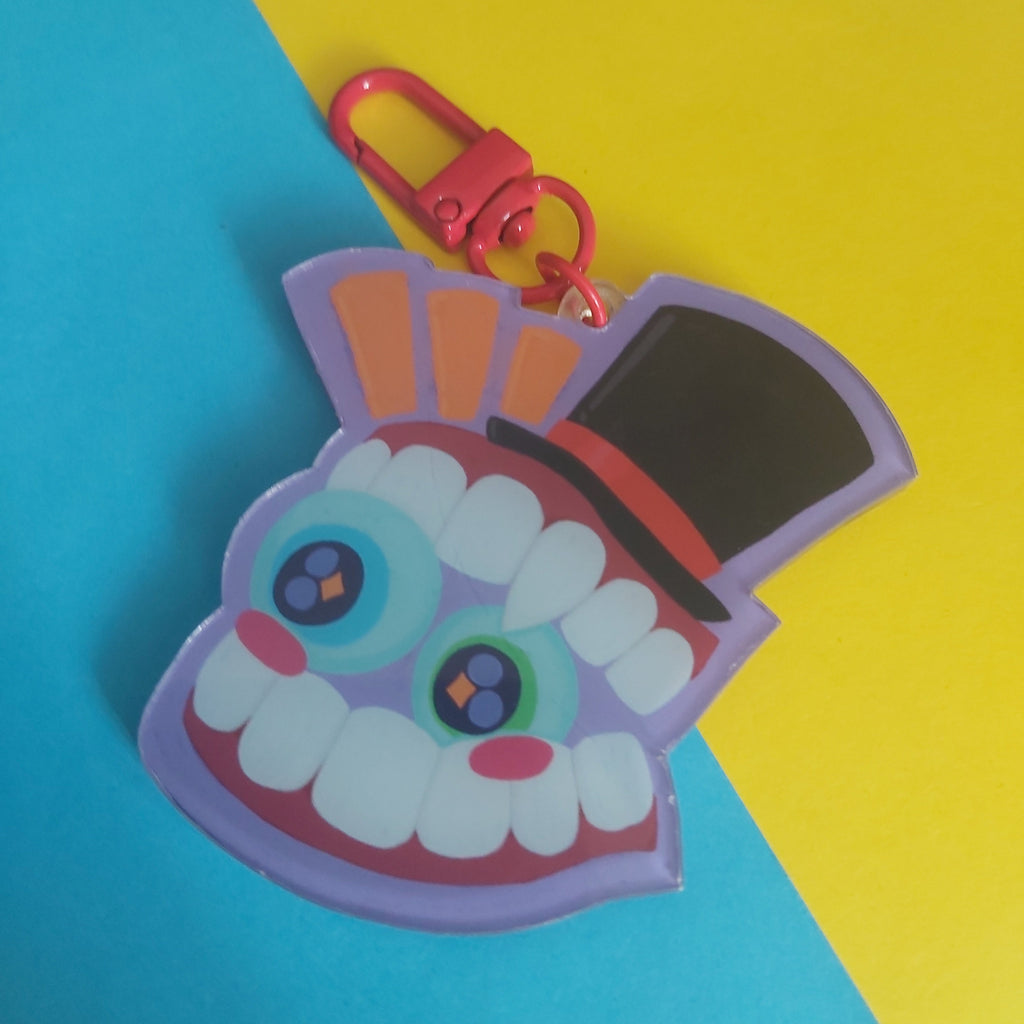A colorful acrylic keychain charm shaped like a stylized monster face wearing a black top hat, attached to a red metal clasp. The charm features white teeth, large blue and purple eyes, pink cheeks, and orange and purple accents, set against a diagonally split yellow and blue background.