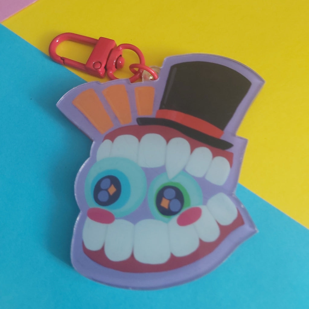 A colorful acrylic keychain charm shaped like a smiling cartoon face with large eyes, white teeth, a black top hat, and a red clasp, photographed on a bright blue and yellow background.