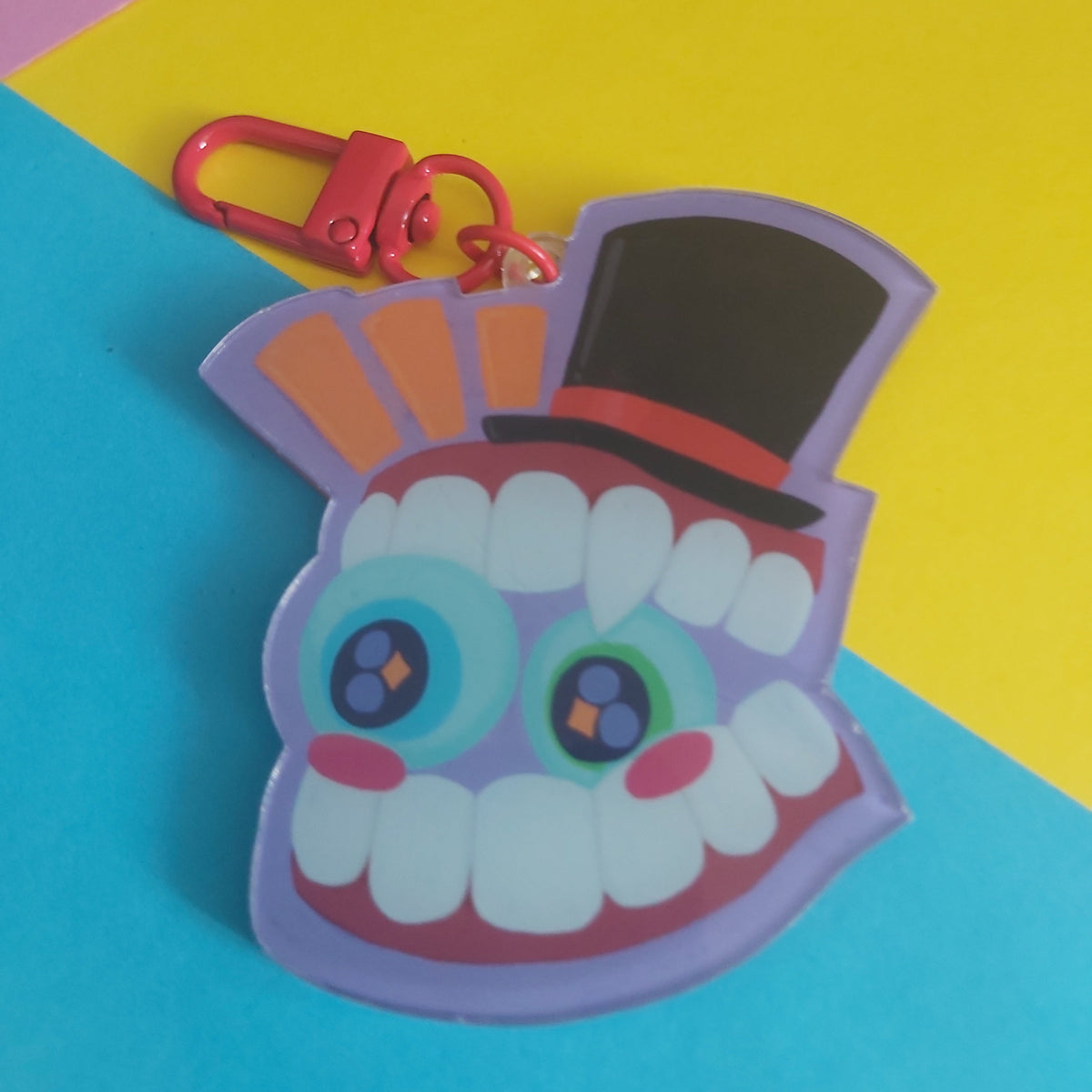 A colorful acrylic keychain charm shaped like a smiling cartoon face with large eyes, white teeth, a black top hat, and a red clasp, photographed on a bright blue and yellow background.