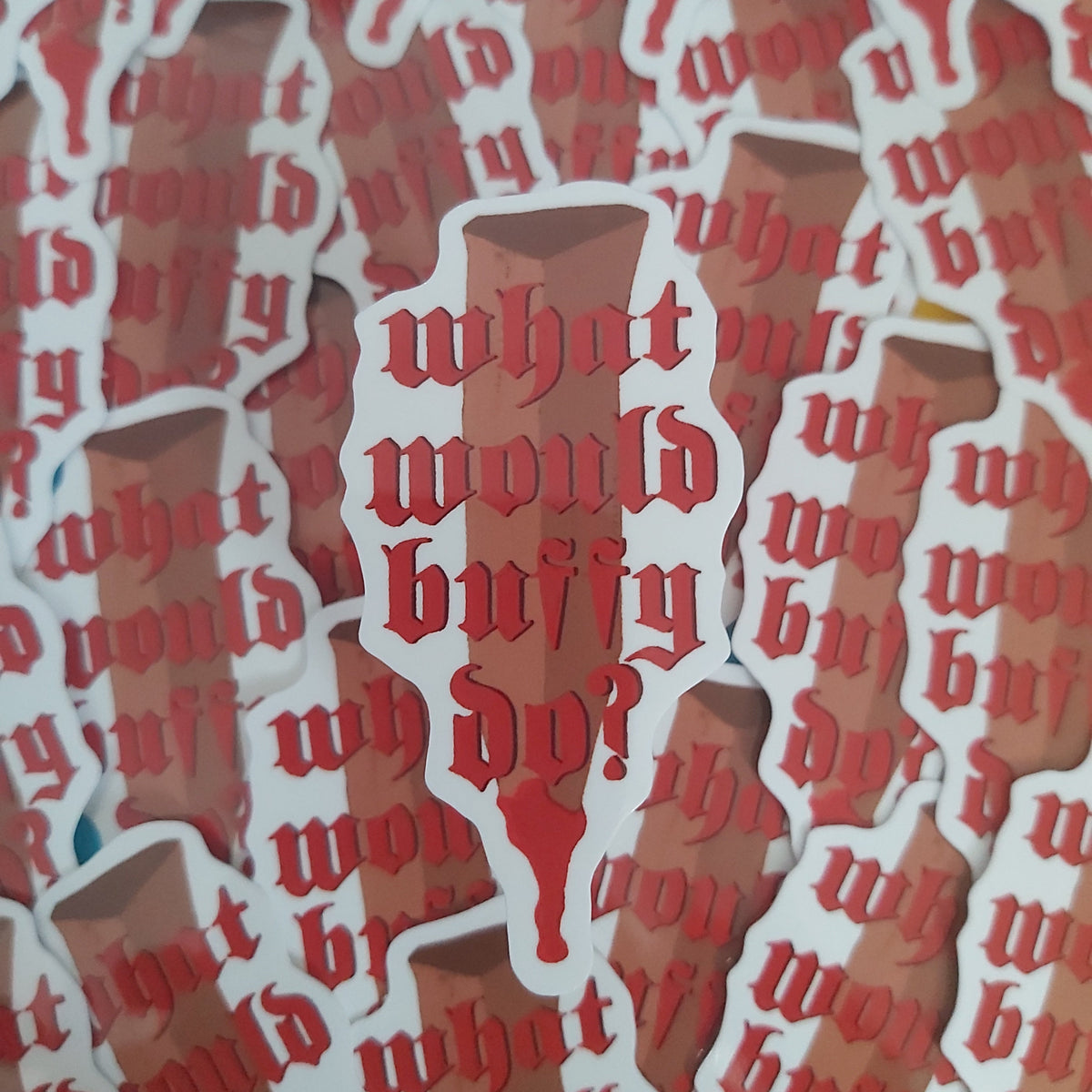 A close-up of multiple die-cut stickers layered on top of each other, featuring the phrase 'what would Buffy do?' in red Gothic-style lettering over a brown silhouette of a wooden stake, with white sticker borders.