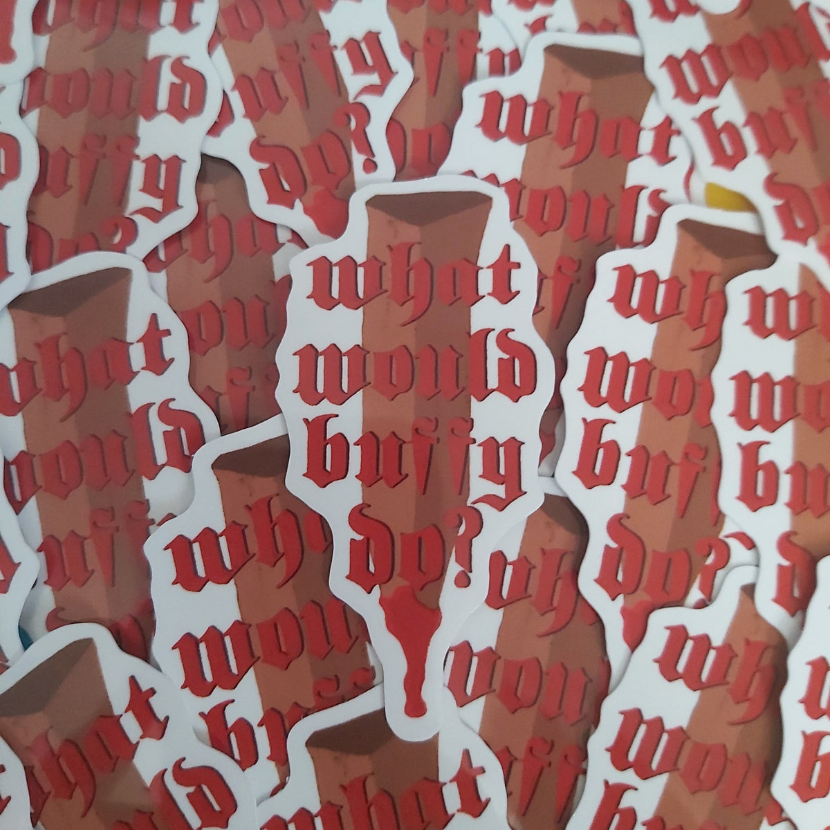 A close-up of multiple die-cut stickers featuring the phrase 'what would buffy do? in red gothic lettering on a white outline, layered over a brown illustrated stake.